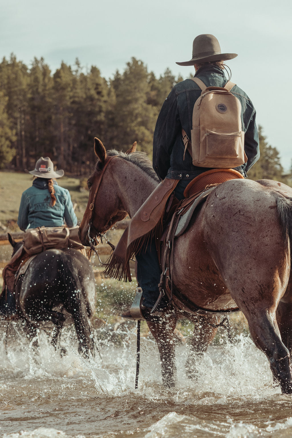 A cowboy rides a brown horse through shallow water in a forested outdoor setting, with another rider visible in the background. The main rider wears a wide-brim hat, dark jacket, and brown leather backpack.