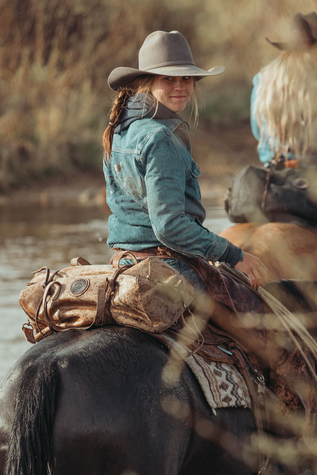 A woman wearing a gray cowboy hat, a distressed teal denim jacket, and a western belt sits on a horse in an outdoor, rustic setting with a custom branded brown leather duffel bag mounted on the back of her horse.