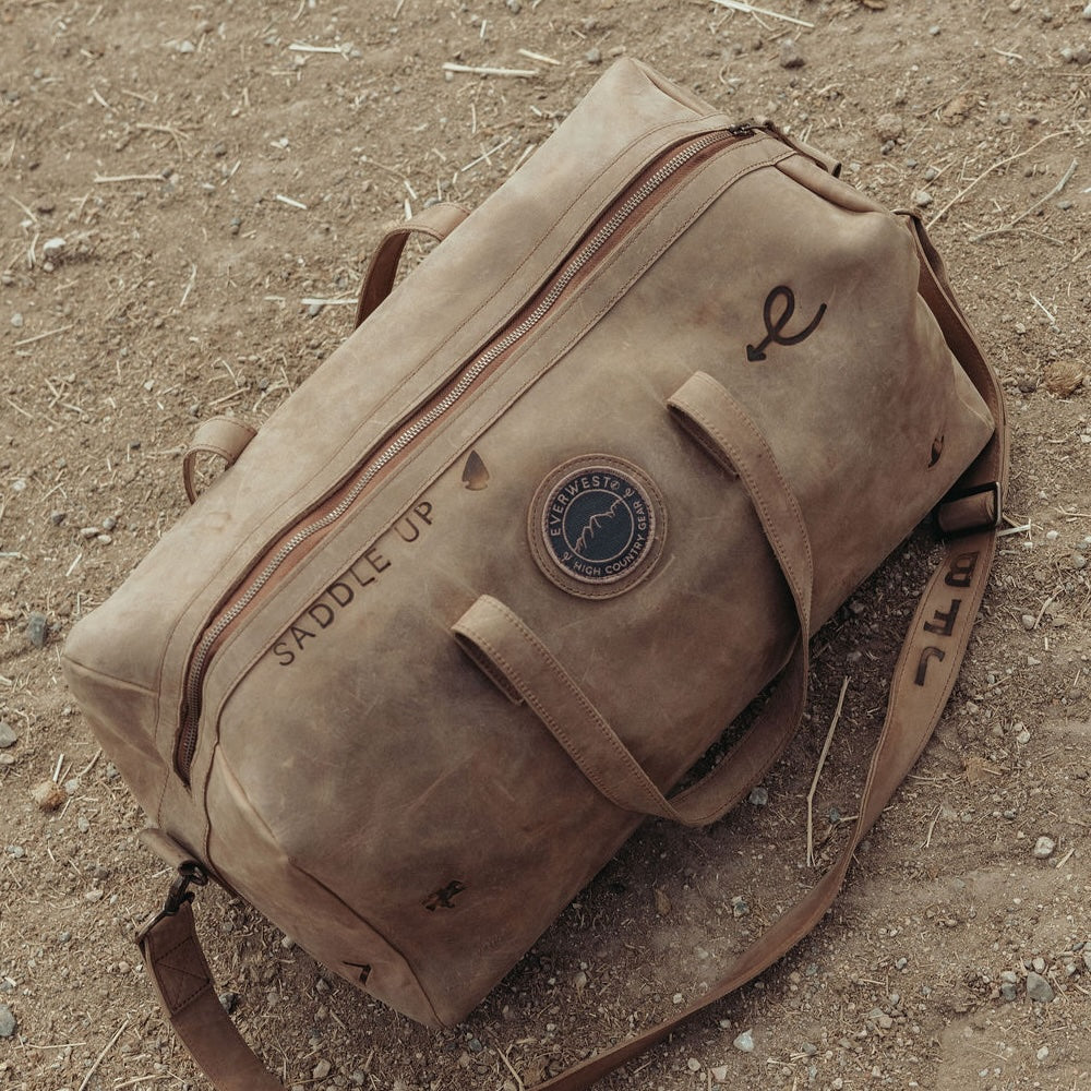 A distressed brown leather duffel bag custom branded with 'Saddle Up' and a few other branding irons, resting on a sandy dirt ground. The bag features a top zipper, leather handles, and a matching shoulder strap custom printed with 'JFB'.
