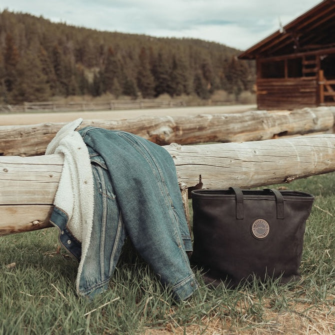 A black leather tote bag with the Everwest logo patch sits on green grass next to a rustic wooden log fence, with a blue denim jacket and a white knit sweater draped over the fence in a country setting with a cabin and pine trees in the background.