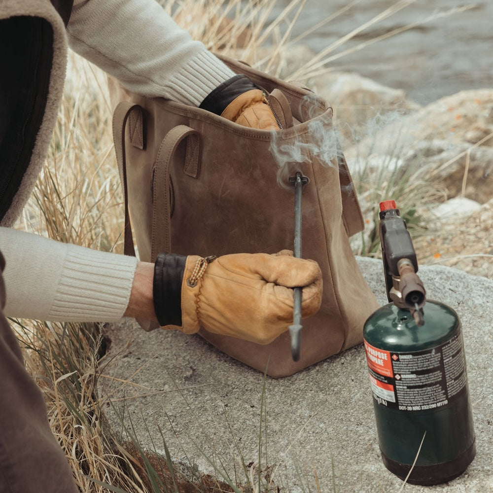 A person wearing tan leather work gloves holds a hot branding iron over a brown leather bag outdoors, with a green propane torch nearby on a stone surface.