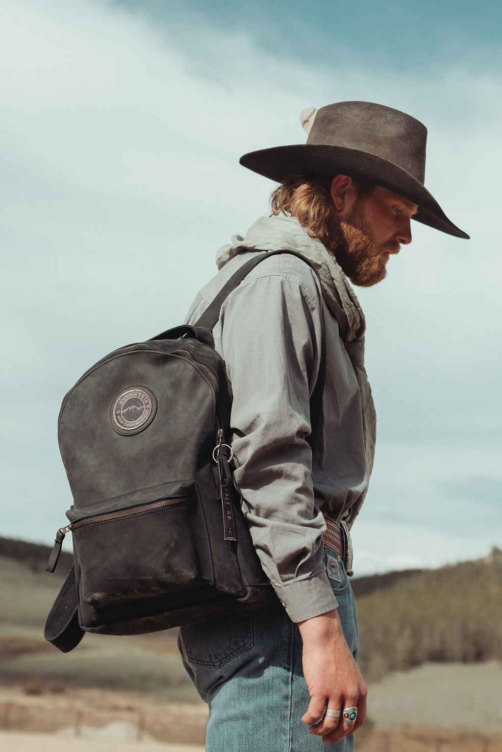 A man in a cowboy hat, gray shirt, and jeans wearing a distressed black leather backpack with the Everwest circular logo patch, standing outdoors against a cloudy sky.