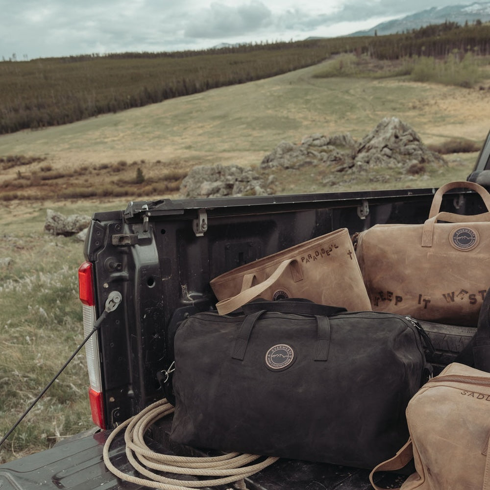 A large black leather duffel bag, a brown leather branded and customized tote, and other matching travel bags loaded into the back of a pickup truck, set against a rugged outdoor landscape with hills and trees.
