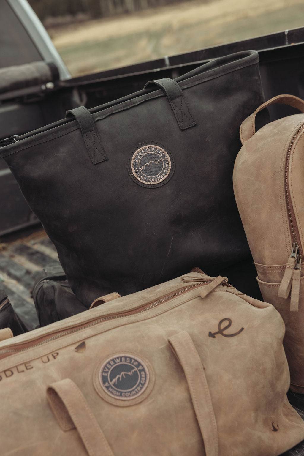 A collection of distressed western leather gear, including a black leather tote bag, a brown leather duffel bag, and a brown leather backpack, arranged together in the bed of a truck. the brown leather duffel has custom brands on it.