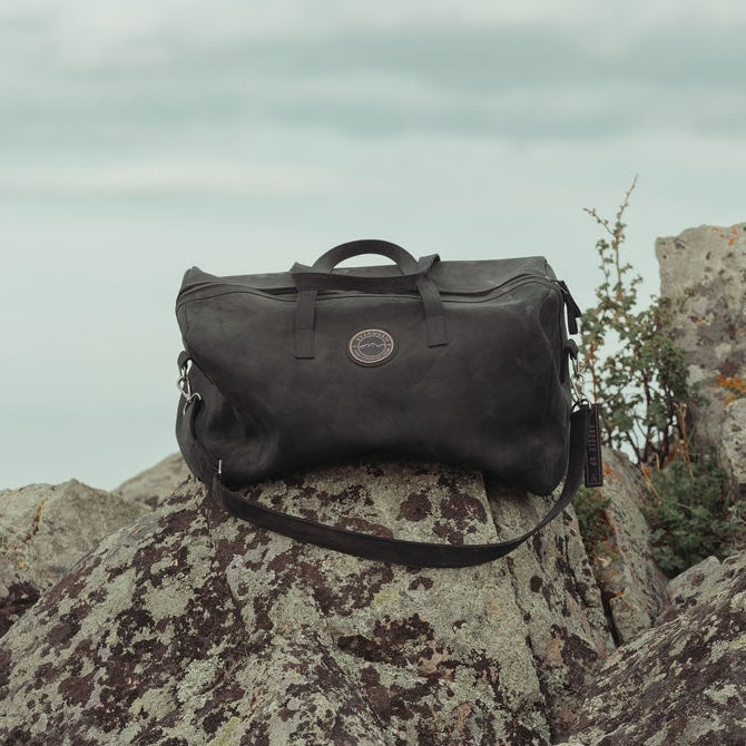 A large black leather duffel bag with the Everwest logo patch, resting on a large mossy rock under an overcast sky.