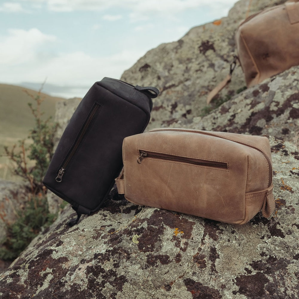 Two leather toiletry bags, one black and one brown, displayed on a large mossy rock outdoors with a blurred natural landscape in the background.