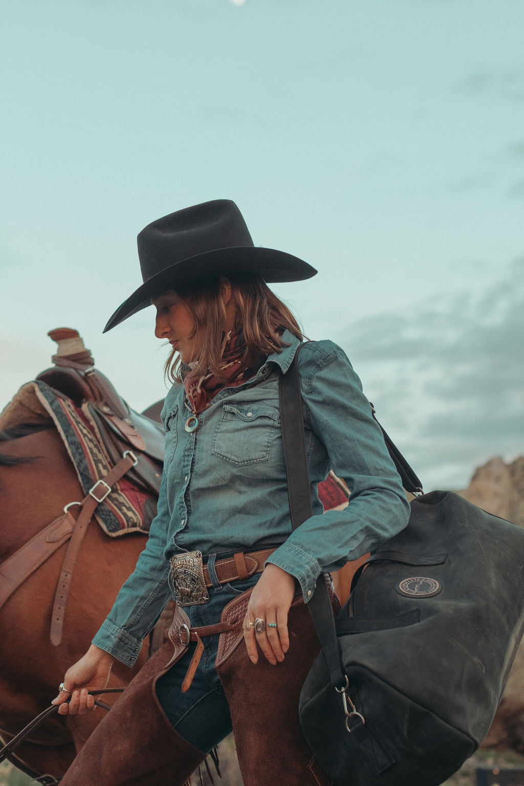 A woman wearing a black cowboy hat, a teal denim shirt, and a brown scarf sits on a brown horse outdoors, carrying a black leather duffel bag.