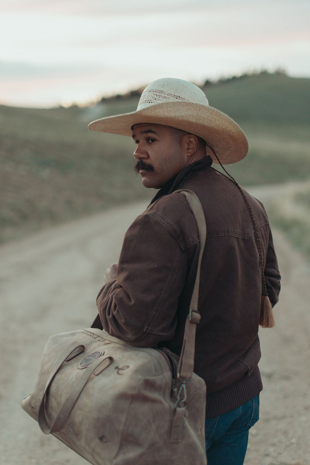 A man wearing a wide-brim straw cowboy hat, brown jacket, and blue jeans stands on a dirt road carrying a large brown leather duffel bag with custom brands on it, over his shoulder, with a blurred outdoor landscape in the background.