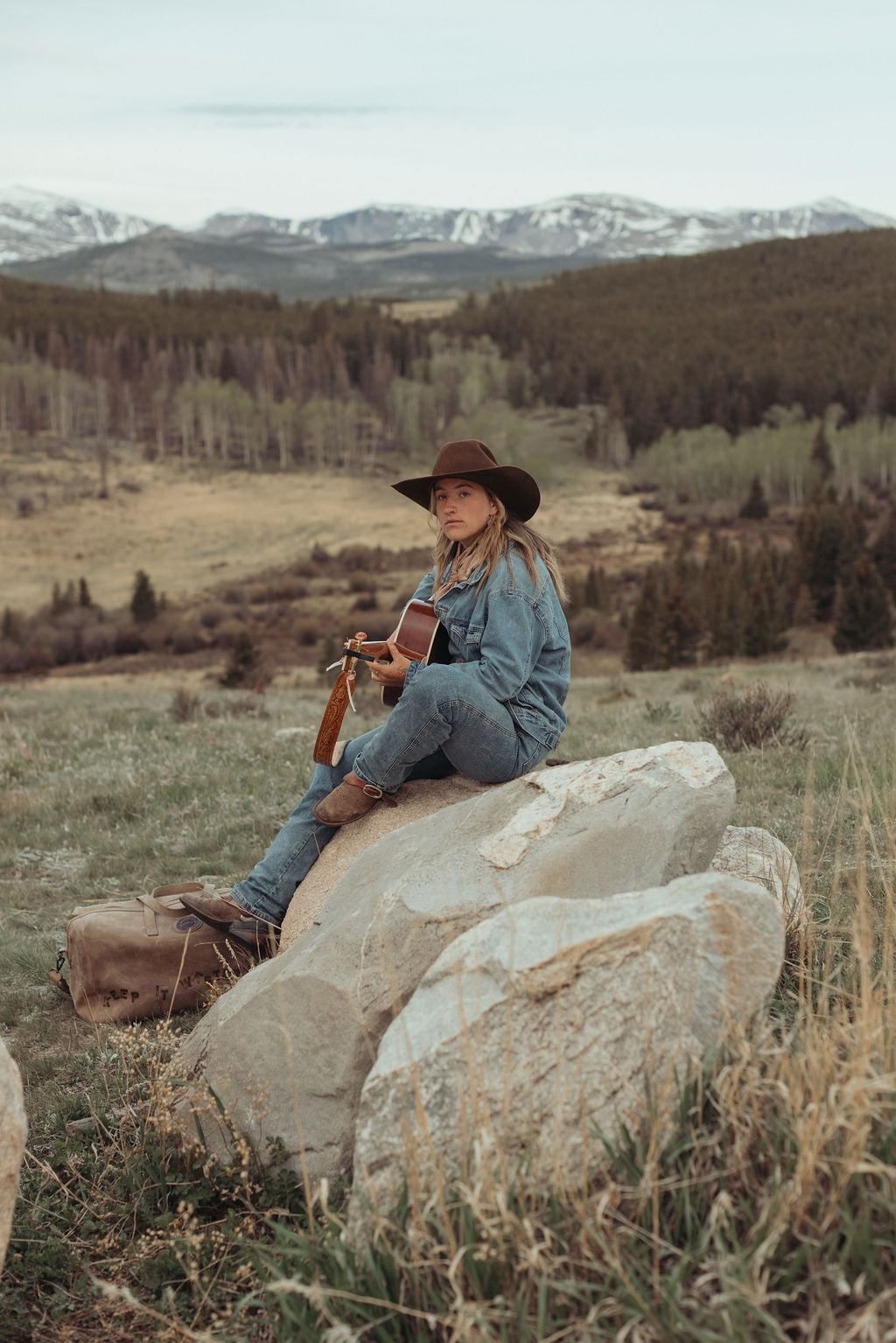 A woman in a blue denim outfit and brown cowboy hat sits on large rocks in a grassy mountain landscape, holding a guitar. Snow-capped mountains and pine trees are visible in the background. a custom branded large brown leather duffel bag sits at her feet.