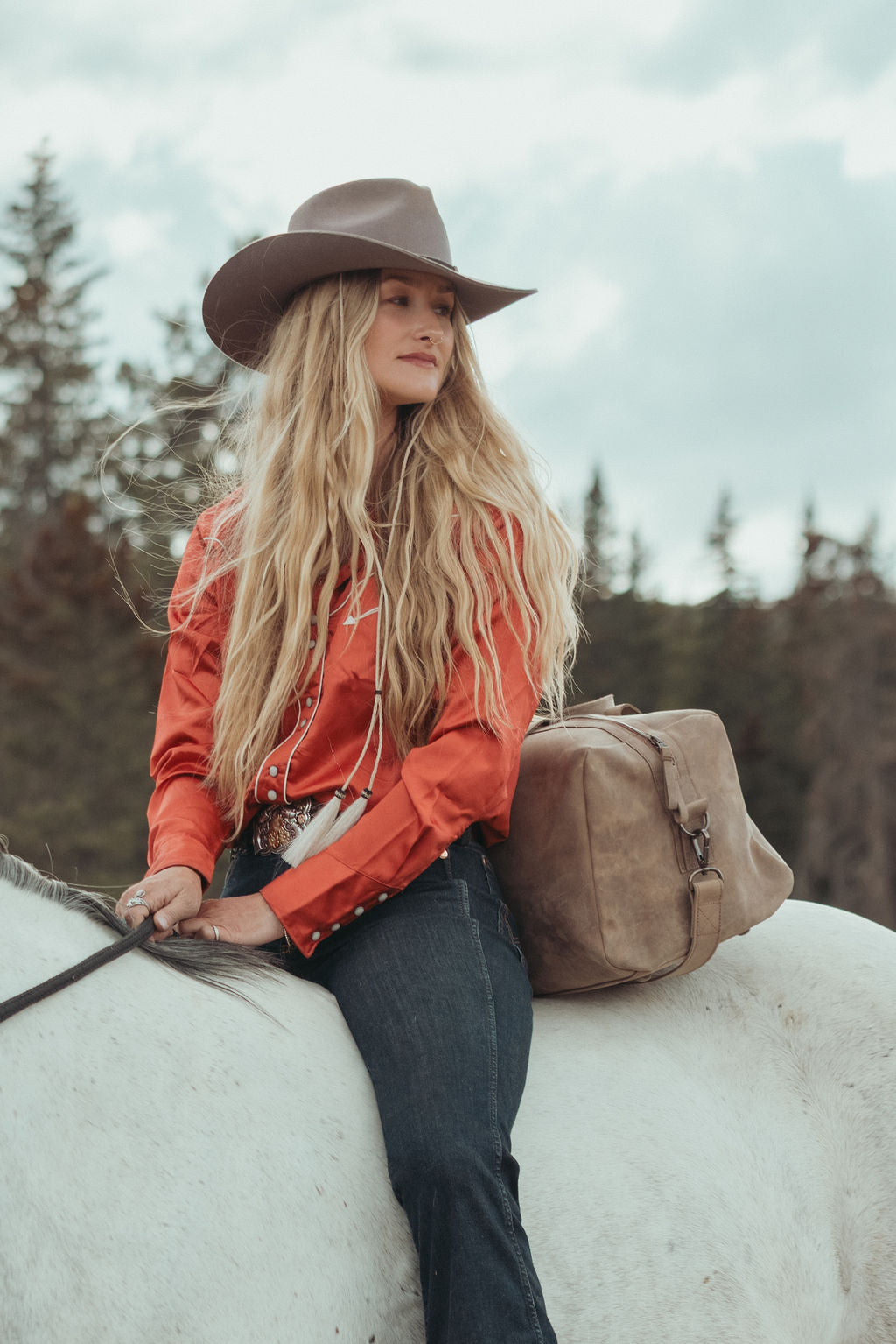 A woman with long blonde hair wearing a brown cowboy hat, rust-orange western shirt, and dark jeans sits on a white horse outdoors. A large brown leather bag rests behind her on the horse, with trees and an overcast sky in the background.