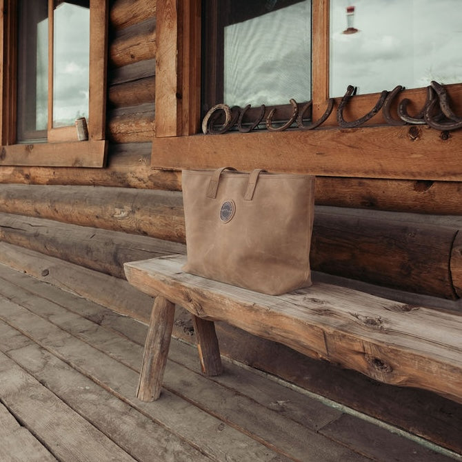 A brown leather tote bag with the Everwest logo patch sits on a rustic wooden bench in front of a log cabin porch.