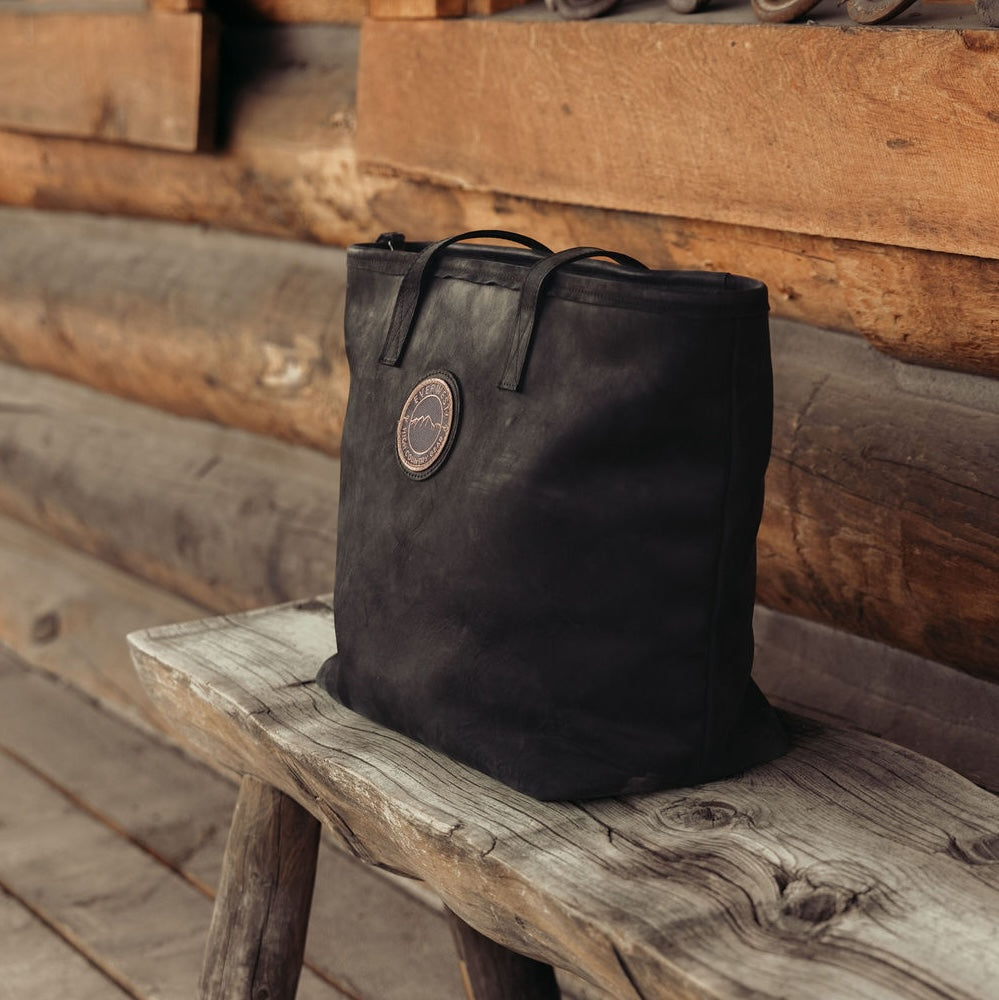 A black leather tote bag with the Everwest logo patch, resting on a rustic wooden bench in front of a log cabin wall.