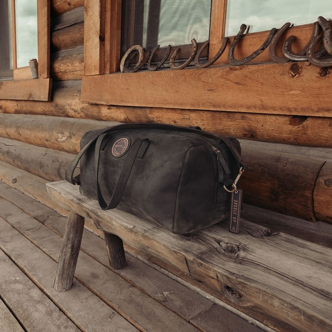A black leather duffel bag with the Everwest logo patch and a hanging tag, resting on a rustic wooden porch bench in front of a log cabin wall.