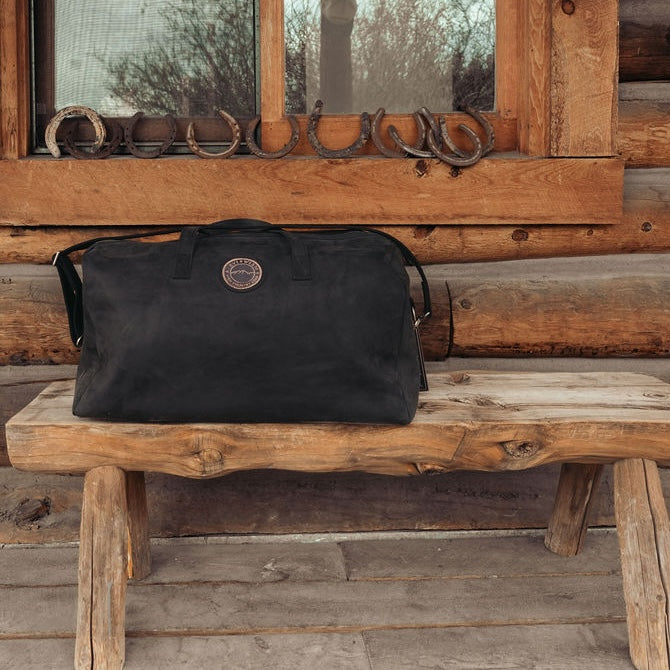 A large black leather duffel bag with the Everwest logo patch sits on a rustic wooden bench in front of a log cabin window with horseshoe decorations.