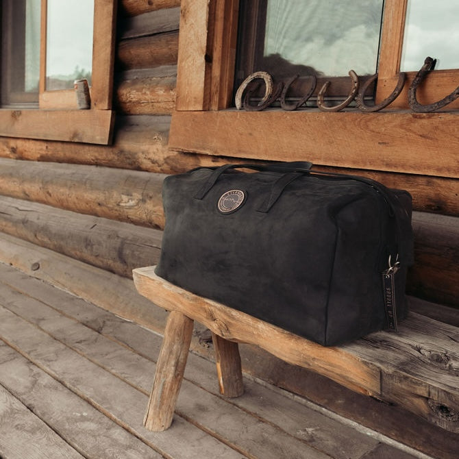 A large black leather duffel bag with the Everwest logo patch sits on a rustic wooden bench in front of a log cabin porch with horseshoe decorations.