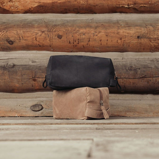 Two leather toiletry bags, one black and one brown, stacked on a rustic wooden log background.