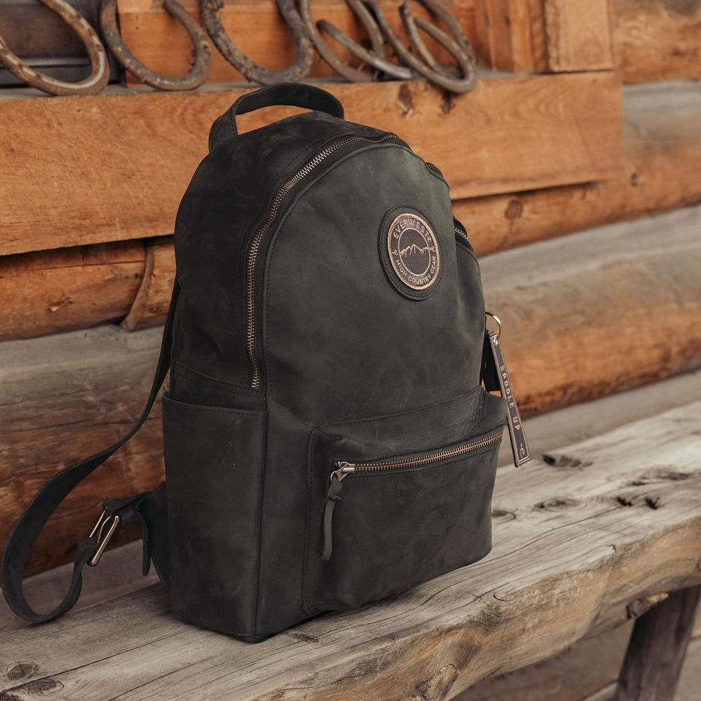 Black leather backpack resting on a wooden bench in front of a wooden log cabin.