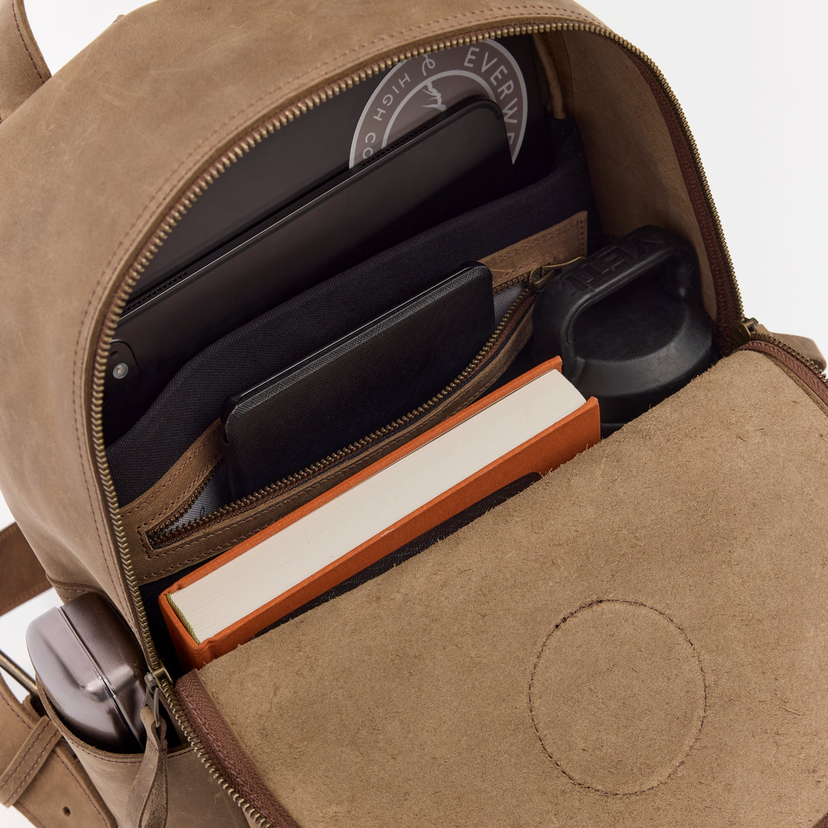 Open brown leather backpack showing electronic devices and a book inside.