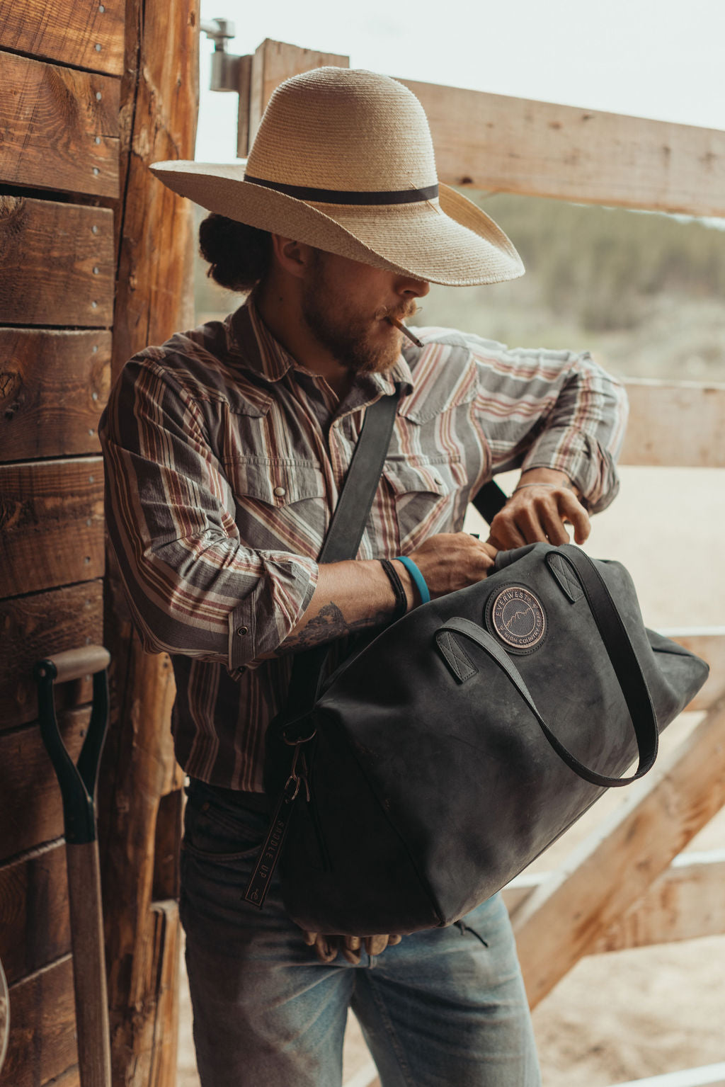 A man in a cowboy hat and striped shirt searches in his duffel bag while wearing a distressed large black leather duffel bag with an Everwest logo patch, set against a rustic wooden background.