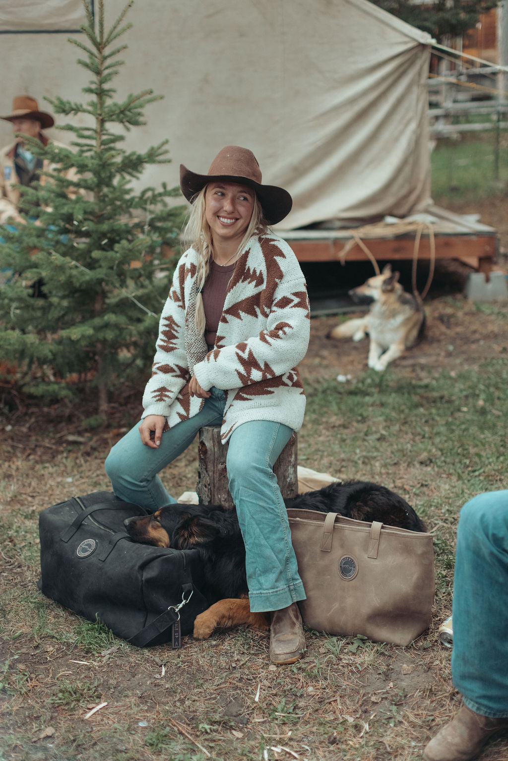 A woman wearing a brown wide-brim hat, a brown and cream geometric patterned cardigan, and teal jeans sits on a tree stump outdoors at a campsite. A distressed black leather duffel bag and a brown tote bag sit on the ground beside her, with dogs resting inside the bags.