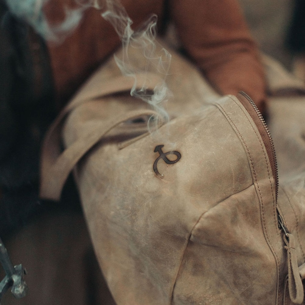 A close-up of a distressed brown leather duffel bag with a custom branded Everwest symbol on the front, partially obscured by smoke, with a person's arm visible in the background.