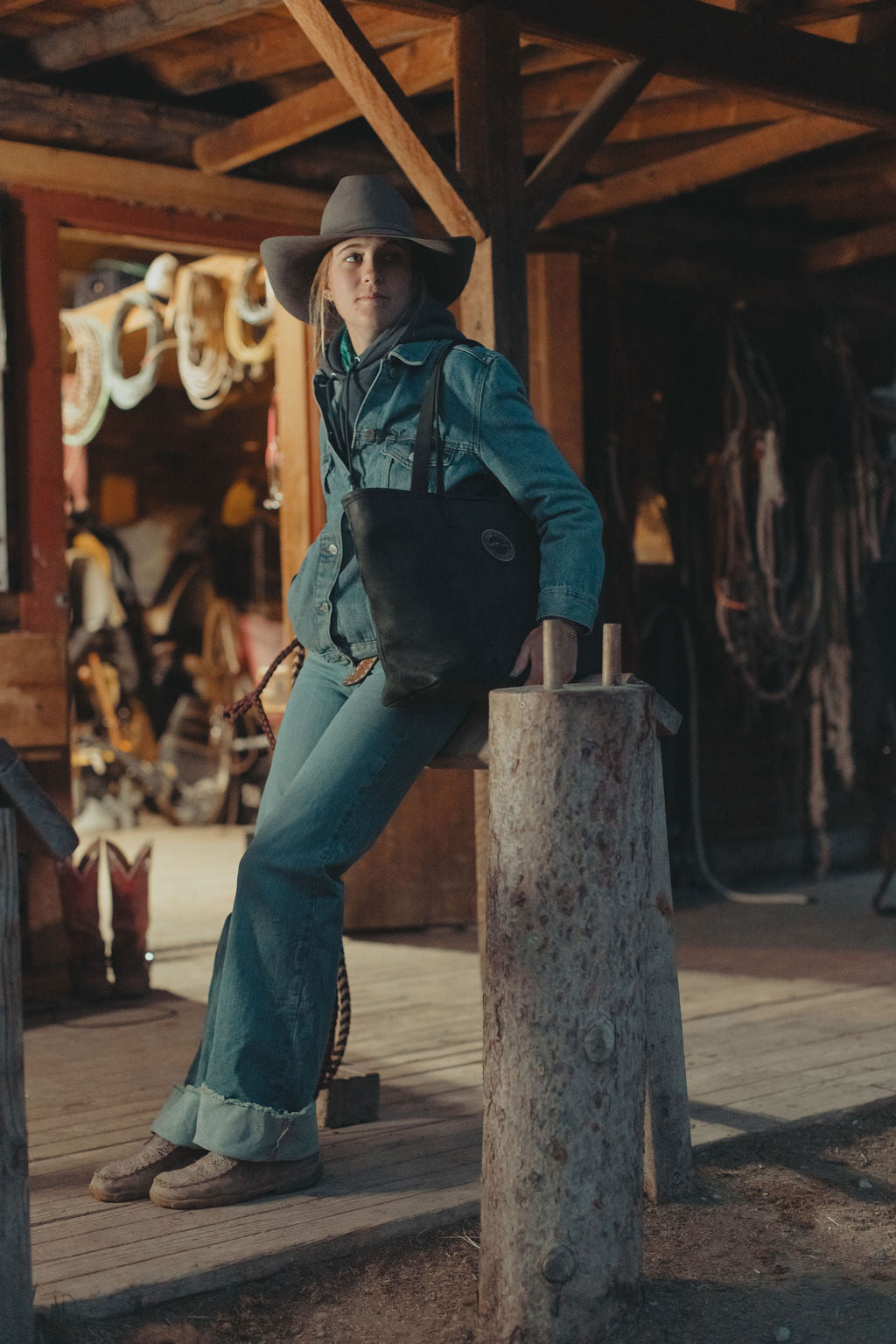 A woman wearing a teal denim western outfit with matching jeans, a cowboy hat, and a black leather tote bag, leaning against a rustic wooden post in a barn setting.