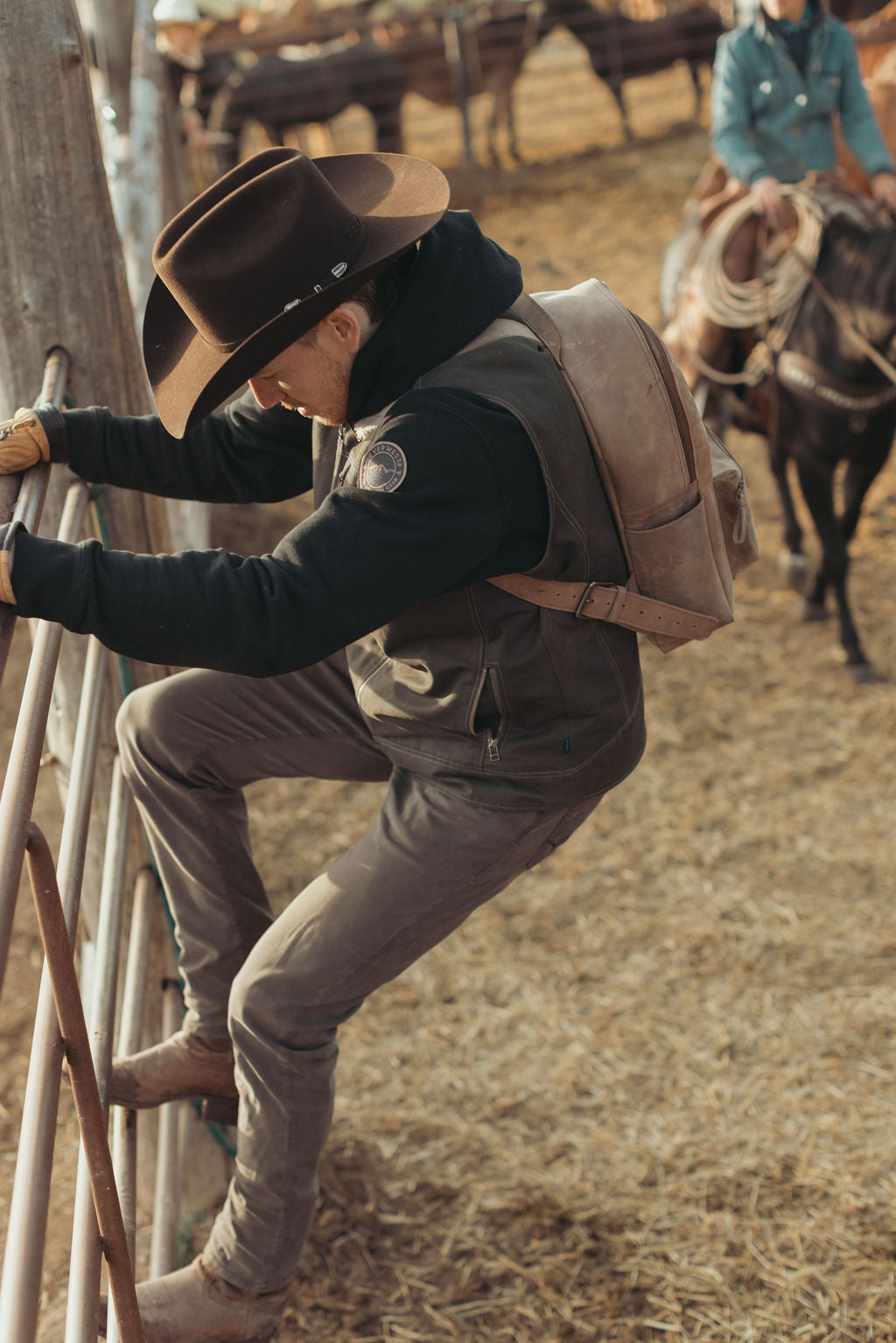 A cowboy in a brown hat, black shirt, and brown vest climbs a metal gate in a dusty ranch arena, with horses and another rider blurred in the background while wearing a brown leather backpack.