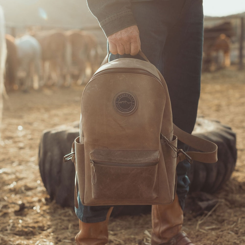 A person in a barn holding a brown leather backpack. The backpack has the Everwest logo patch on the upper front, a zippered front pocket, and side pockets, with a warm, backlit atmosphere and blurred cows in the background.