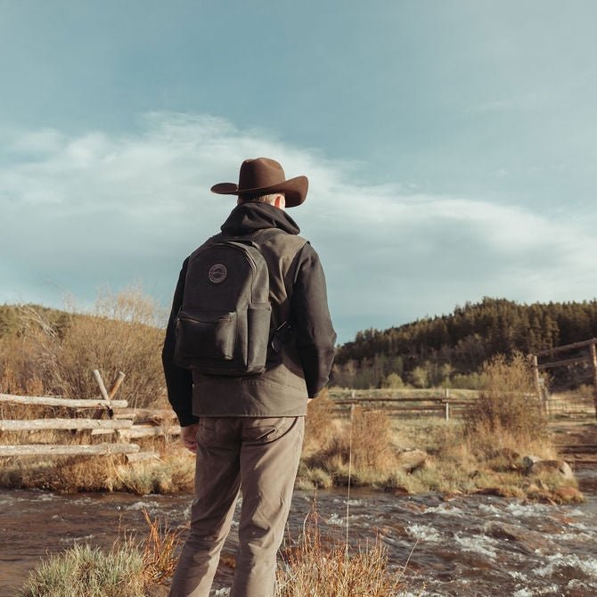 A man wearing a brown cowboy hat, dark jacket, and khaki pants stands with his back to the camera, carrying a black leather backpack while standing near a flowing stream in an outdoor rural landscape.