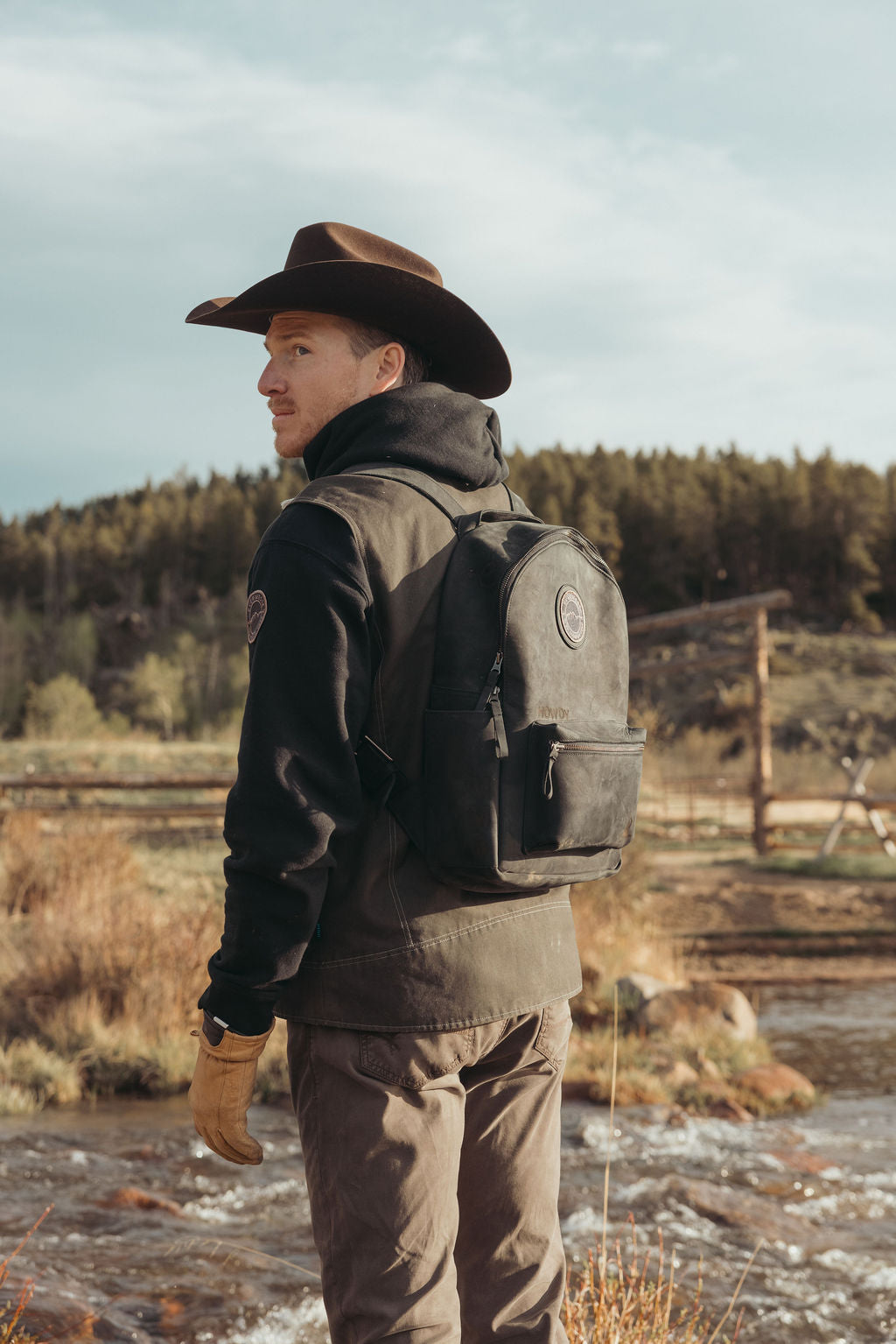 A man wearing a brown cowboy hat, black hoodie, olive green vest, tan pants, and a black leather backpack stands in a dry outdoor landscape near a stream.