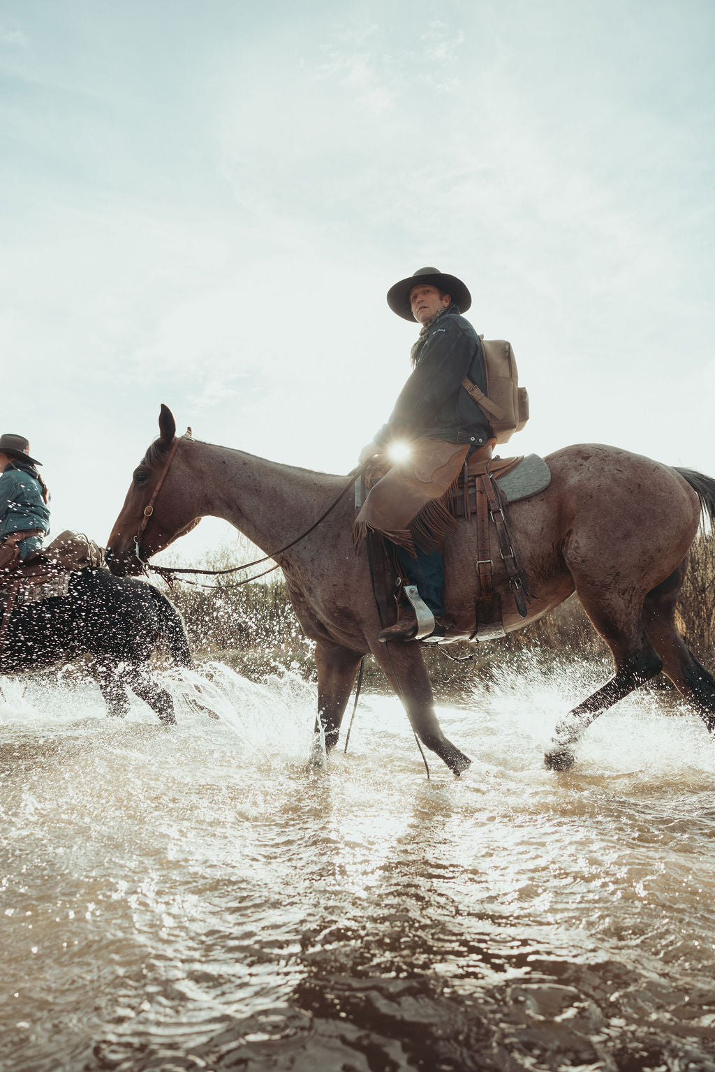 A person riding a brown horse through shallow water, with another rider and horse partially visible in the background. The rider wears a dark hat, dark top, and light pants, and a brown leather backpack, the scene is backlit by bright sunlight.