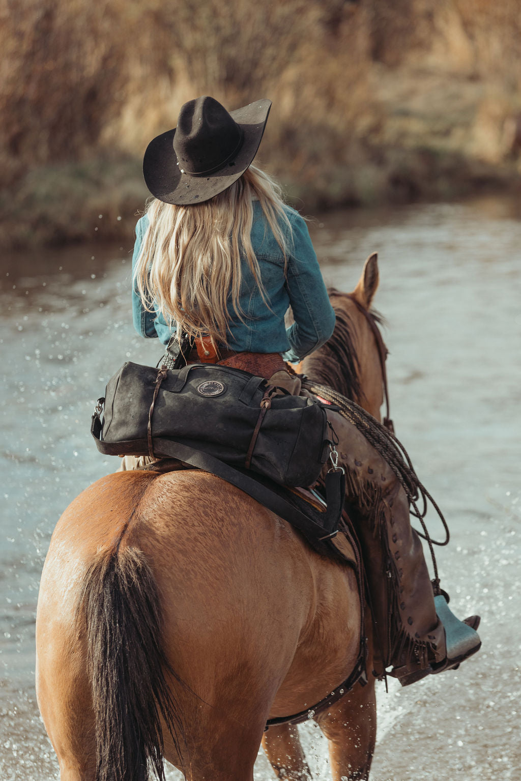 A woman seen from behind riding a brown horse through shallow water, wearing a black cowboy hat, turquoise top, and tall brown boots. A black leather duffel bag sits on the back of the horse.