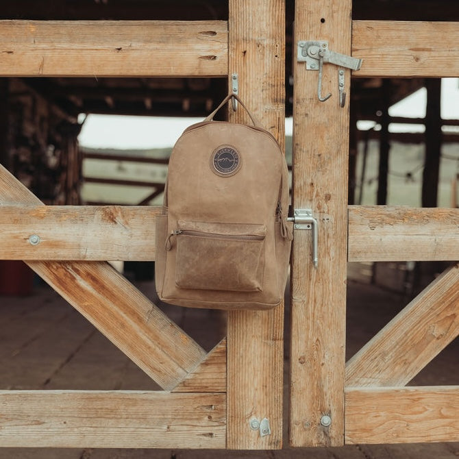 A distressed brown leather backpack hangs from a metal hook on a rustic wooden barn door.