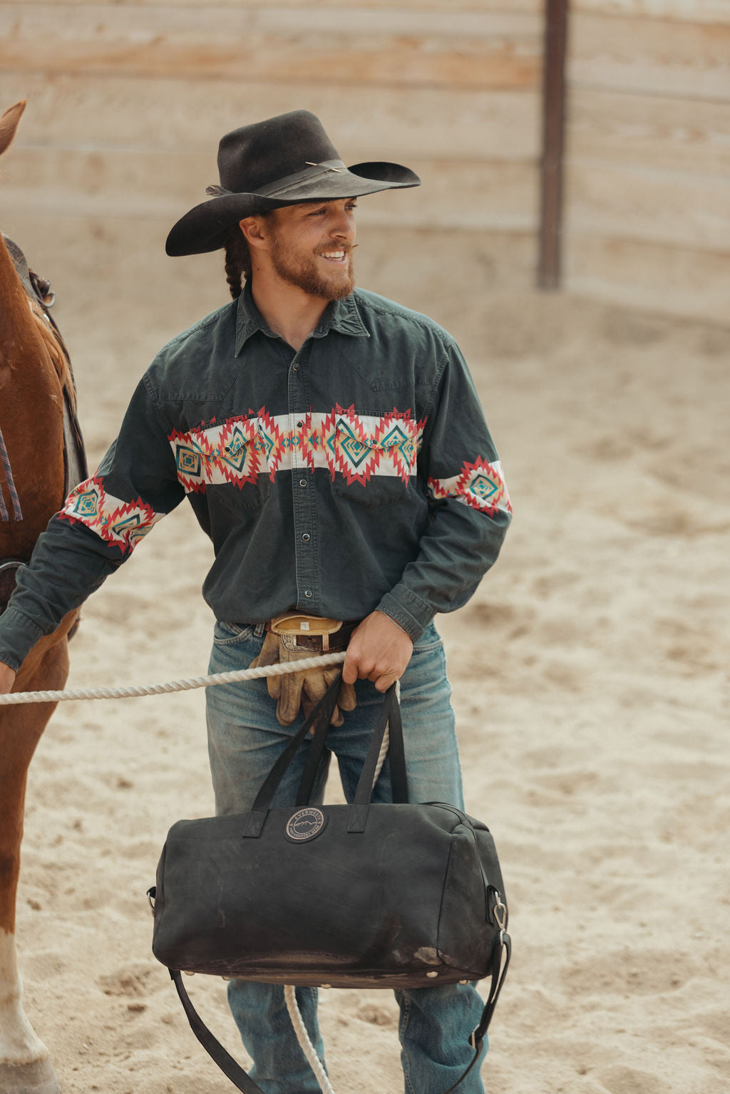 A cowboy wearing a black hat and a dark green long-sleeve shirt with a colorful geometric pattern across the chest and sleeves, holding a distressed black leather duffel bag in a sandy arena next to a horse.