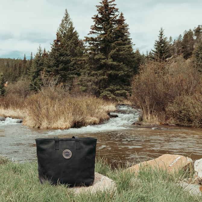 A black leather tote bag with the Everwestlogo patch sits on a stone in a grassy area next to a flowing river, with pine trees in the background.