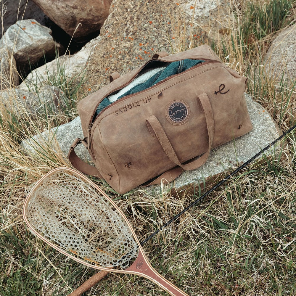 A distressed brown leather duffel bag custom branded with 'SADDLE UP' sits open on a stone in a grassy outdoor setting, with a fishing net and rod placed in front of it.