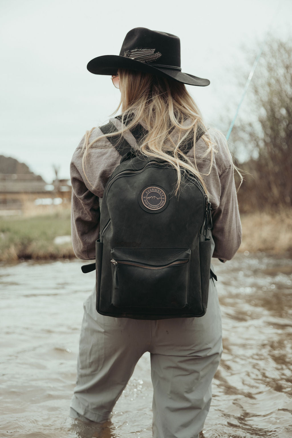 A woman seen from behind standing in shallow water, wearing a black wide-brim hat, a brown jacket, light gray pants, and a black leather backpack with the Everwest circular logo patch on the back.