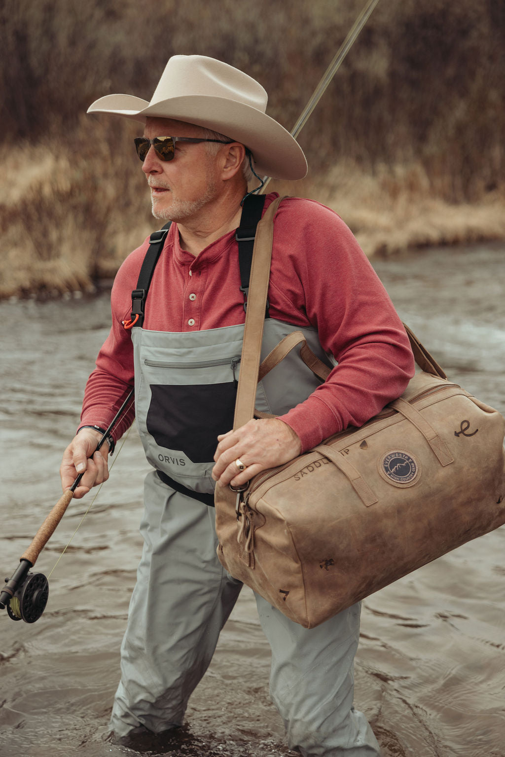 A man wearing a cowboy hat, sunglasses, a red shirt, and gray waders stands in shallow water while carrying a brown leather duffel bag with custom brands on it over his shoulder and holding a fishing rod.