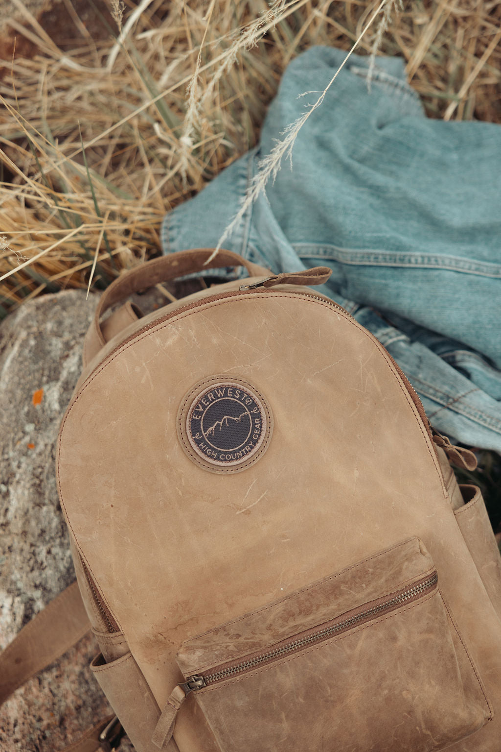 A distressed brown leather backpack with the Everwest logo patch, styled outdoors on a rock with dry grass and a blue denim jacket in the background.