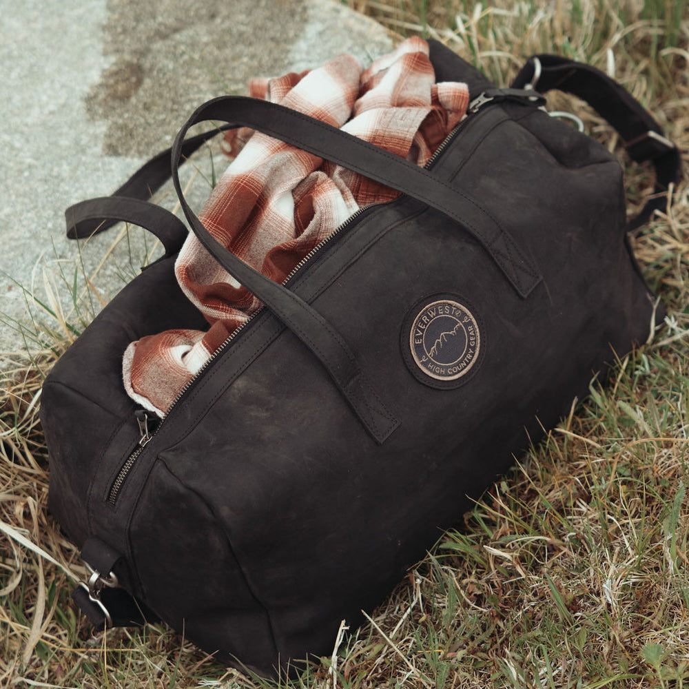 A black leather duffel bag lying on grass outdoors, with a rust-orange plaid blanket stuffed inside. The Everwest brand patch is visible on the side of the bag.