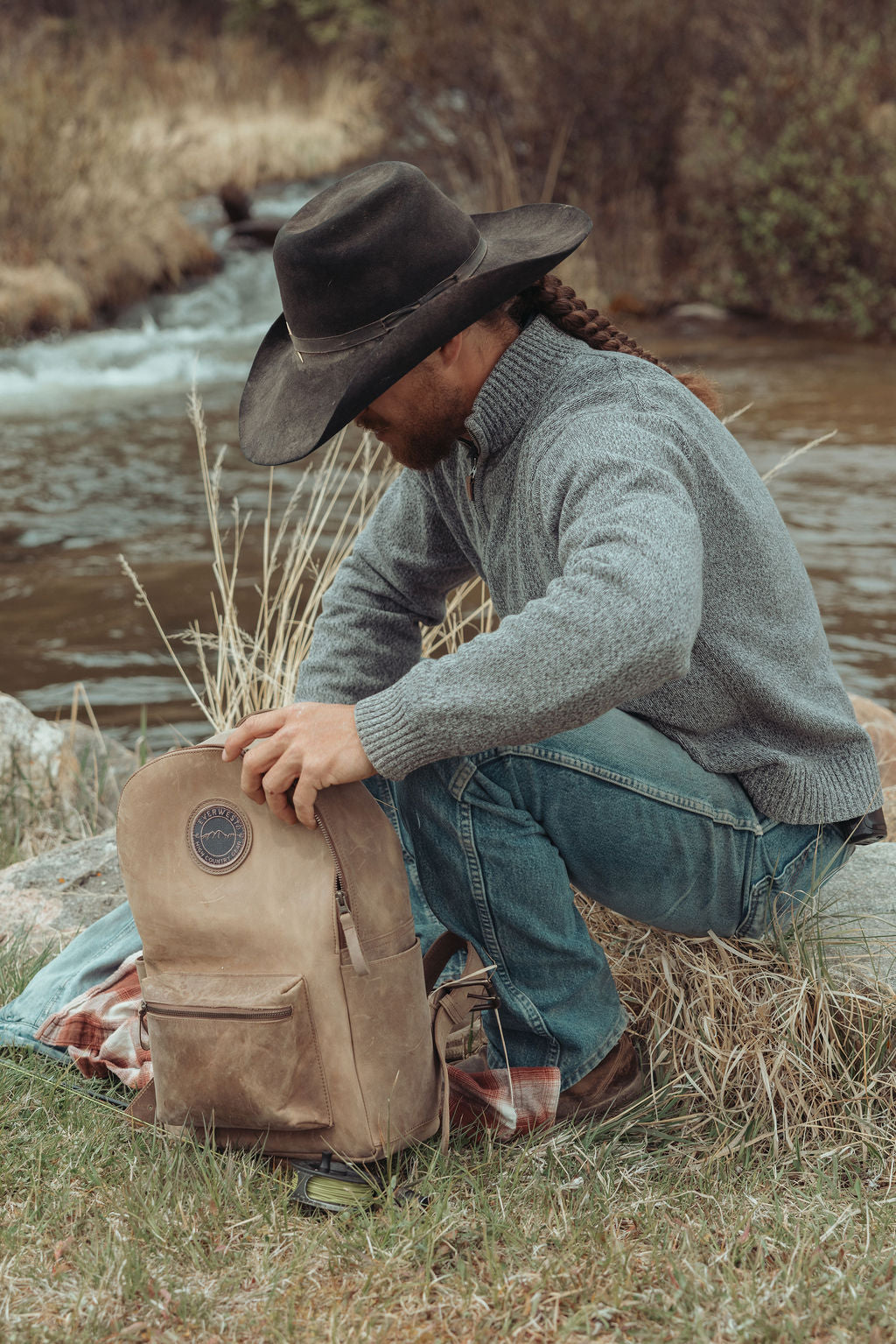 A man in a cowboy hat, gray sweater, and jeans crouches by a stream outdoors, holding a brown leather backpack with the Everwest logo patch.