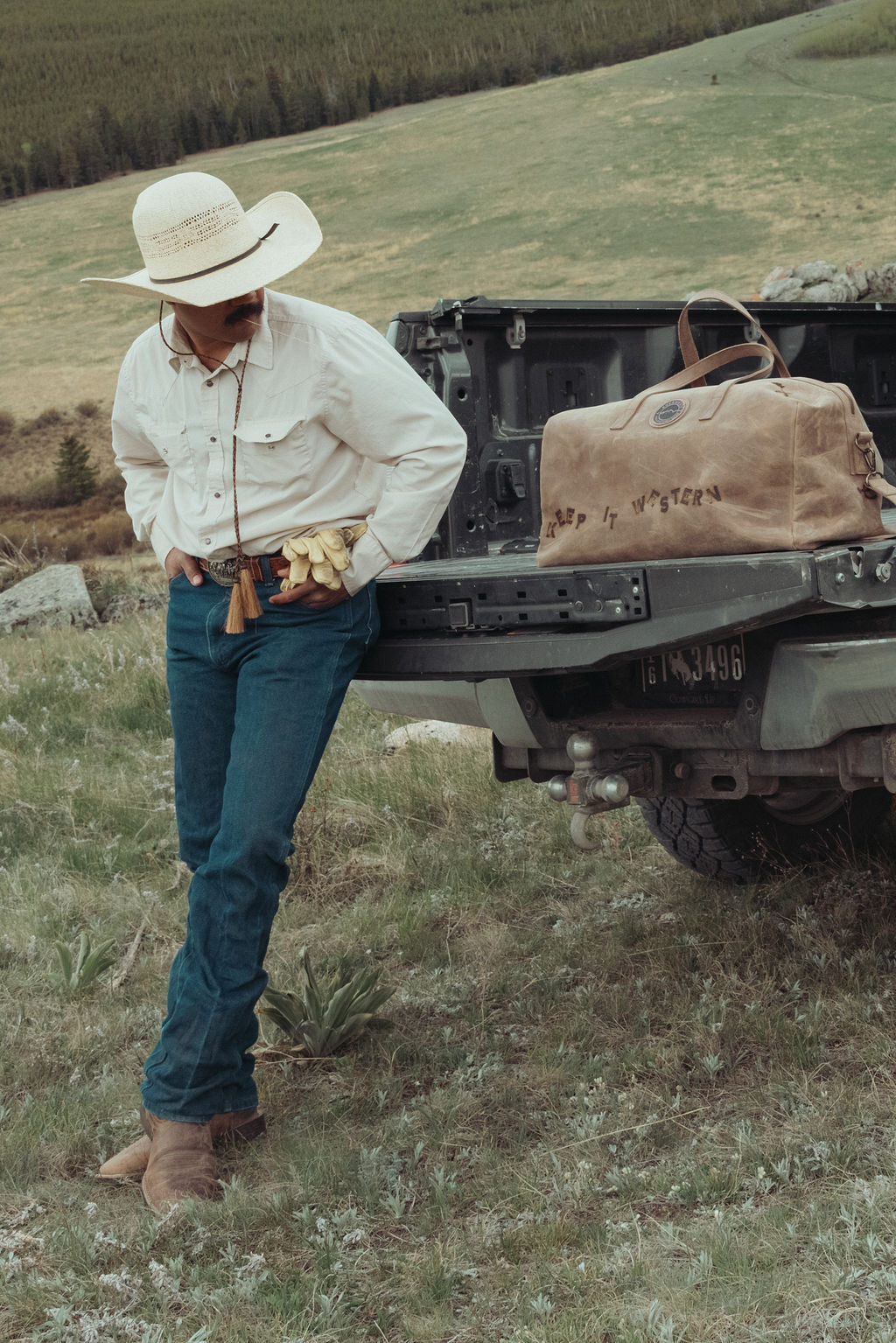 A man in a cowboy hat, cream shirt, and blue jeans stands beside the tailgate of a truck in an outdoor grassy landscape. A distressed large brown leather duffel bag rests in the truck bed with custom brands burned into it.