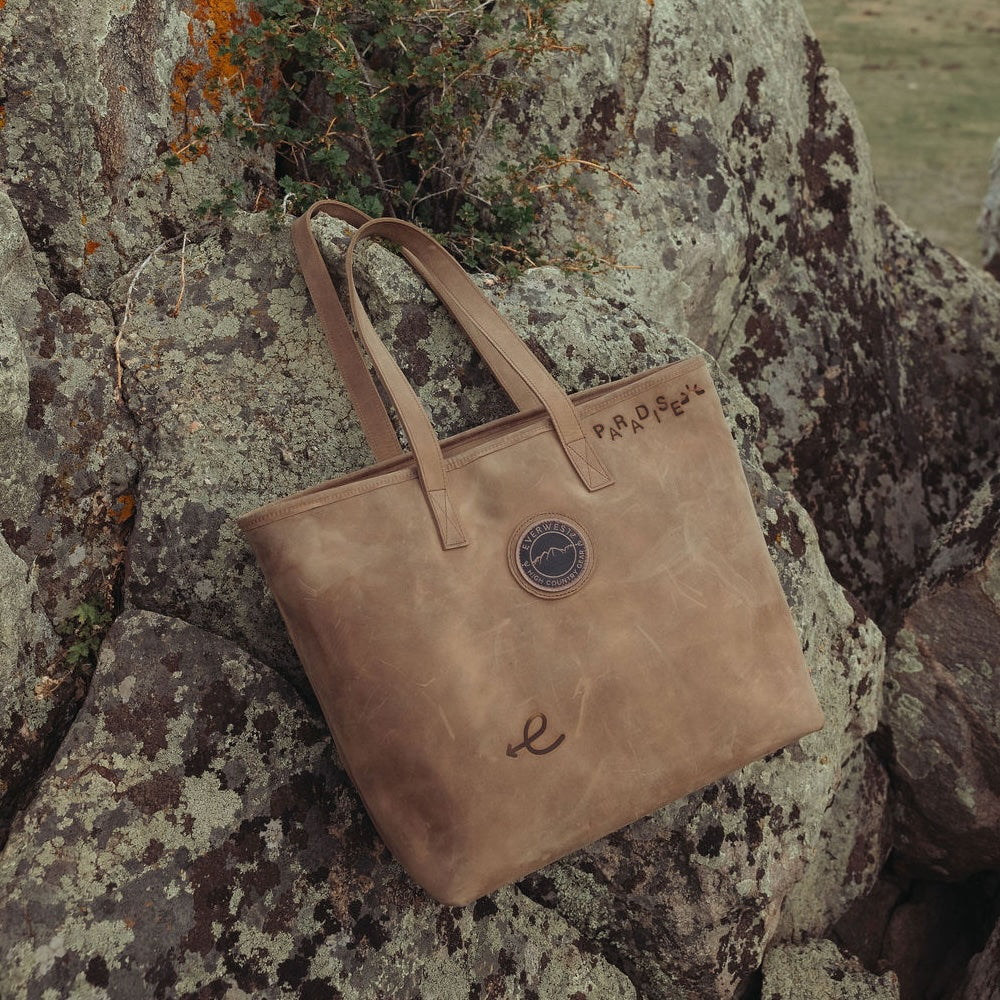 A distressed brown leather tote bag with double shoulder straps, resting against large mossy rocks outdoors. The bag features the Everwest logo patch on the front, a branded cursive script Everwest logo near the bottom, and 'PARADISE' branded on the upper right side.