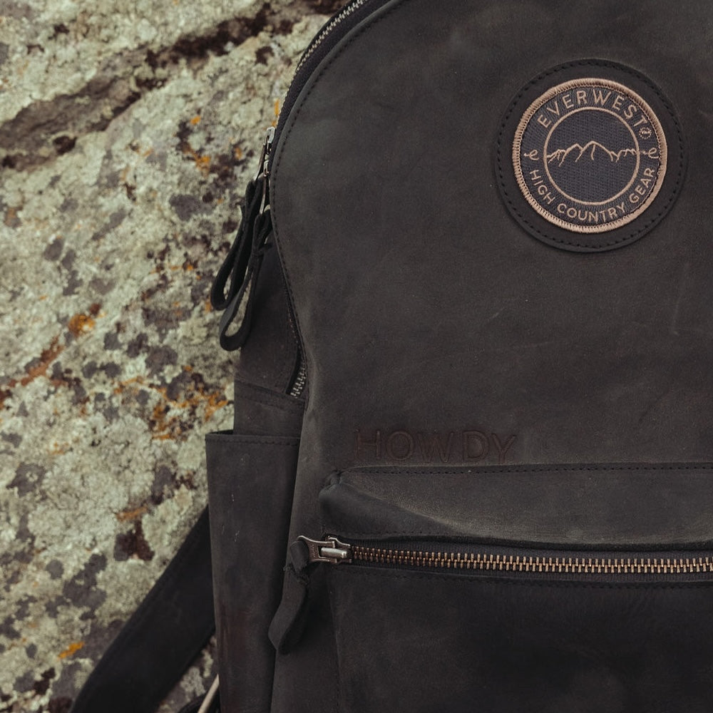 A close-up of a black leather backpack resting against a textured, mossy rock surface. The backpack features a circular 'Everwest High Country Gear' patch on the upper right and a custom branded 'HOWDY' on the front pocket.