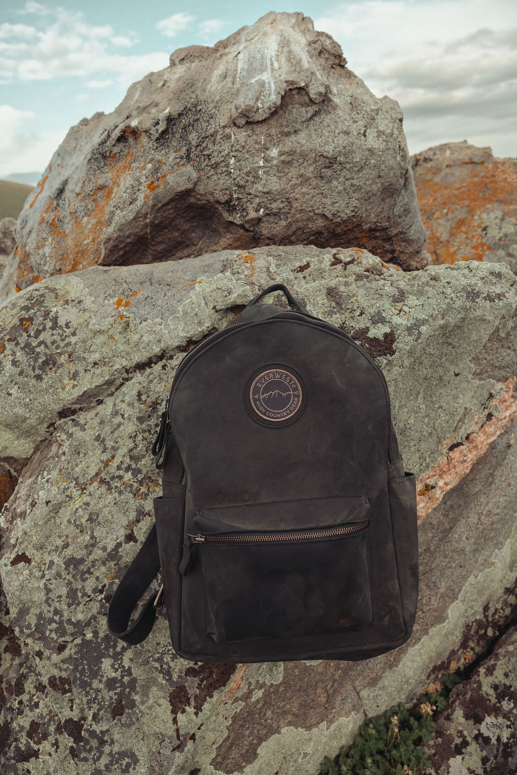 A custom branded black leather backpack with the Everwest logo patch on the front, resting on large, textured gray rocks outdoors under a cloudy sky.