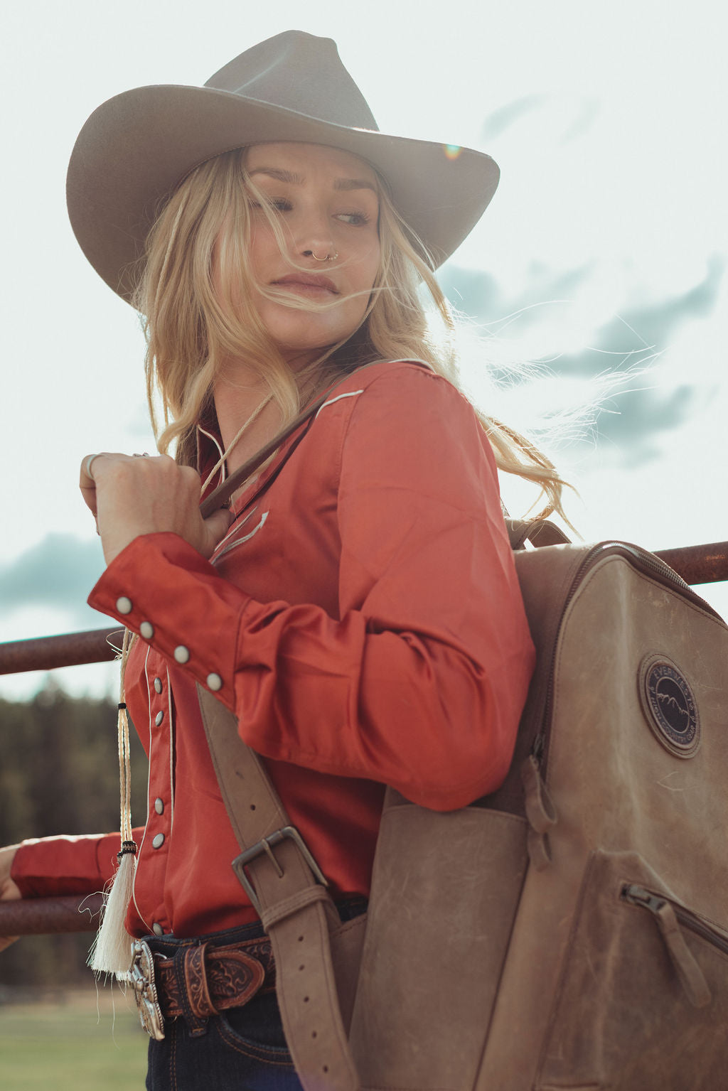 A woman wearing a brown cowboy hat, rust-orange western shirt, and a brown leather backpack, standing outdoors near a fence.