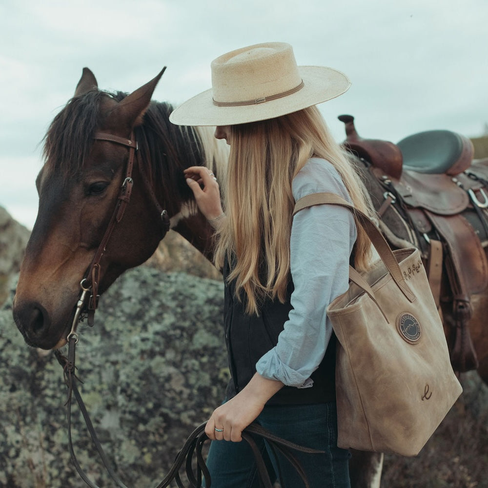 A woman in western attire, including a wide-brimmed straw hat, a light blue shirt, and a black vest, stands next to a brown horse outdoors. She is carrying a brown leather tote bag with the Everwest logo patch and custom brands on the bag.
