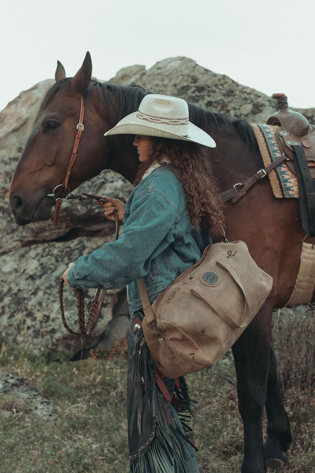 A woman in western attire, including a wide-brimmed hat, denim jacket, and fringed skirt, stands beside a saddled brown horse outdoors against rocky hills. She is carrying a distressed brown leather duffel bag with custom brands on it.
