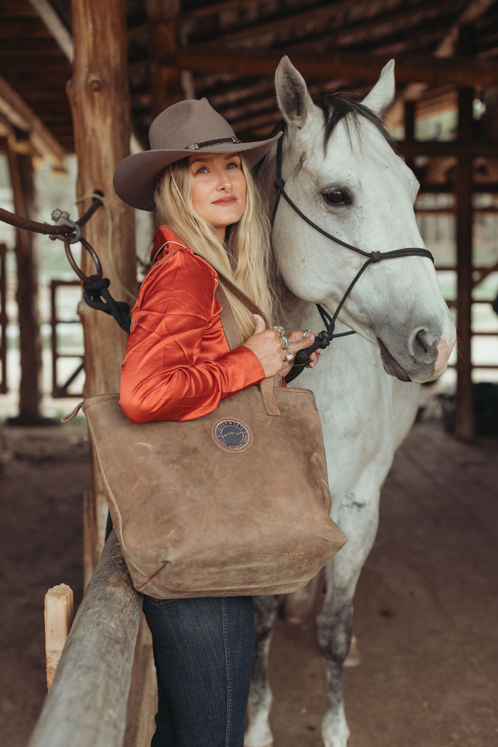 A woman in a cowboy hat and rust-orange jacket stands beside a white horse in a rustic barn, holding a large brown leather tote bag with the Everwest logo patch.