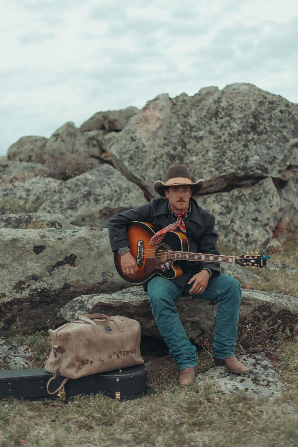 A man in a cowboy hat and dark jacket sits on a large rock outdoors, holding an acoustic guitar. A custom branded, distressed, large brown leather duffel bag and a black hard case rest on the ground nearby, with rocky terrain and an overcast sky in the background.